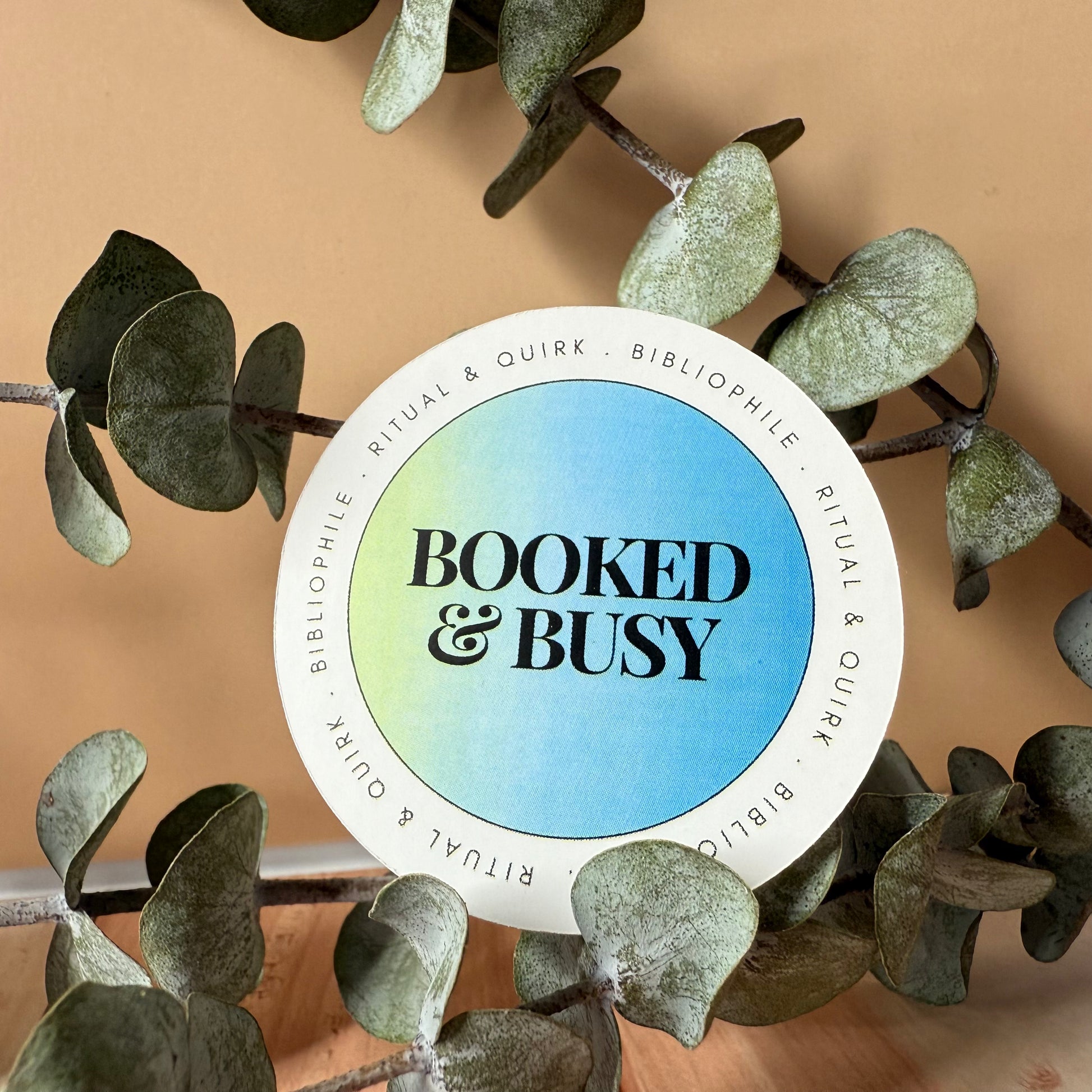 Circular sticker for reading challenge journal with 'Booked & Busy' text surrounded by eucalyptus leaves on a beige background